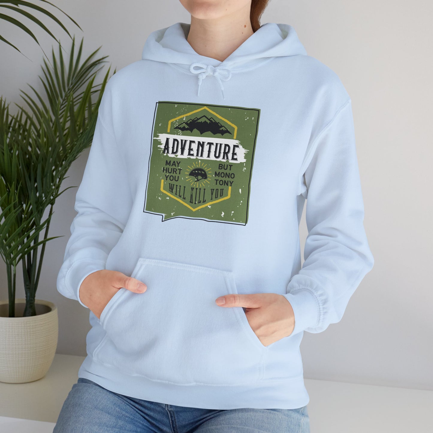 A person wearing a light blue unisex hooded sweatshirt with a motivational quote 'Adventure May Hurt You But Monotony Will Kill You' printed on the front.