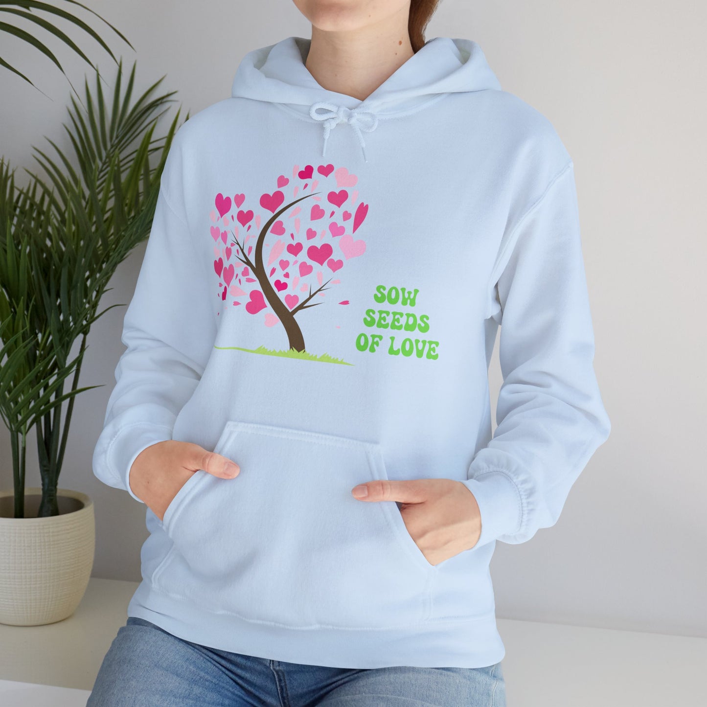 A person wearing a blue hooded sweatshirt with a 'Sow Seeds Of Love' design, featuring a tree with pink leaves and hearts.