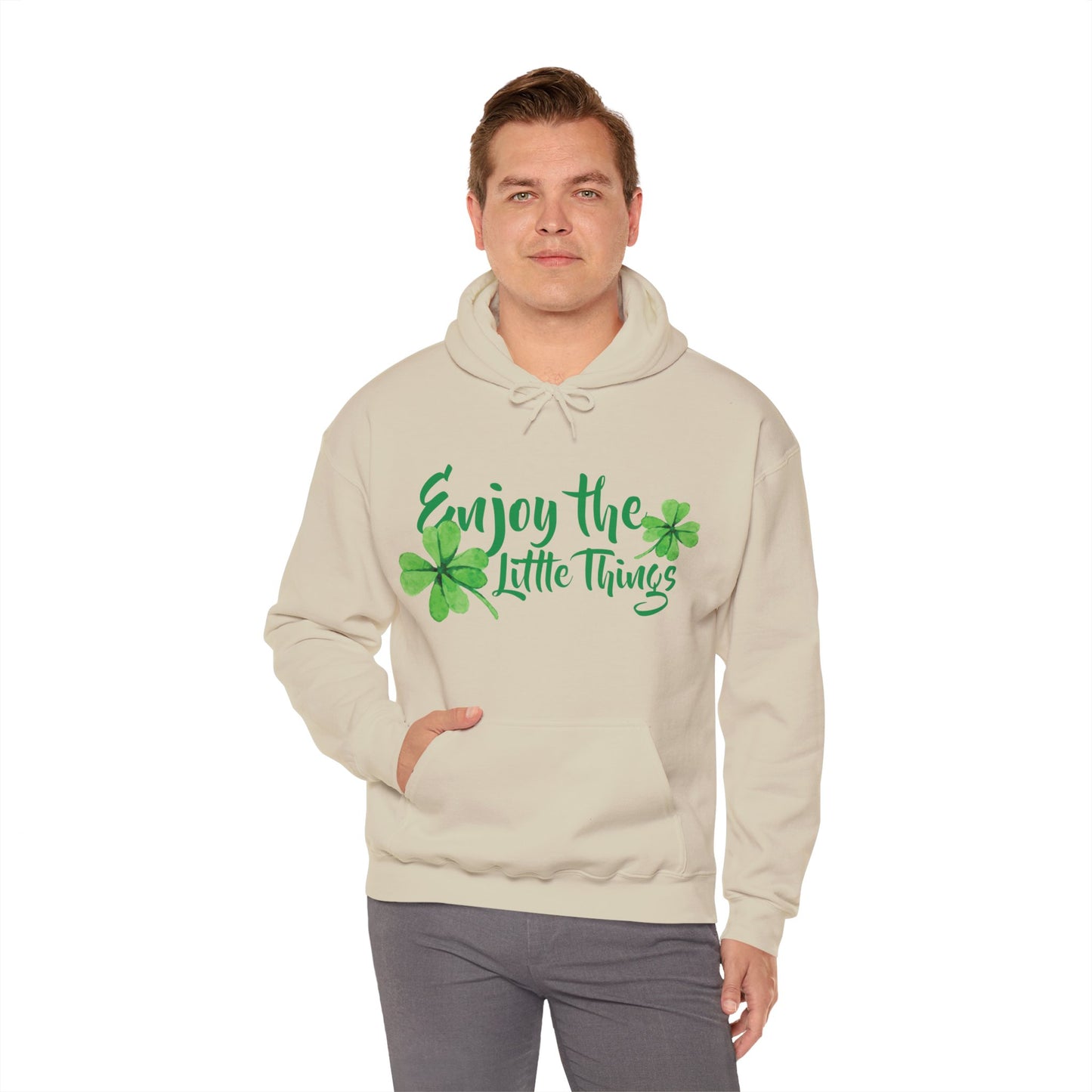 A person wearing a beige unisex hooded sweatshirt with 'Enjoy The Little Things' text and a shamrock graphic design.