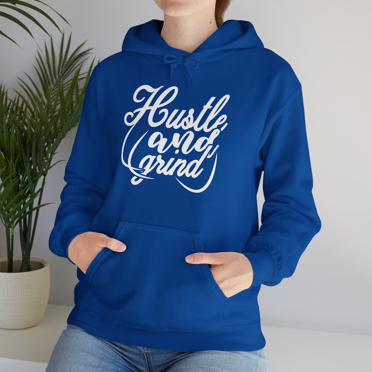 A person wearing a blue unisex hooded sweatshirt with 'HUSTLE AND GRIND' text design on the front.